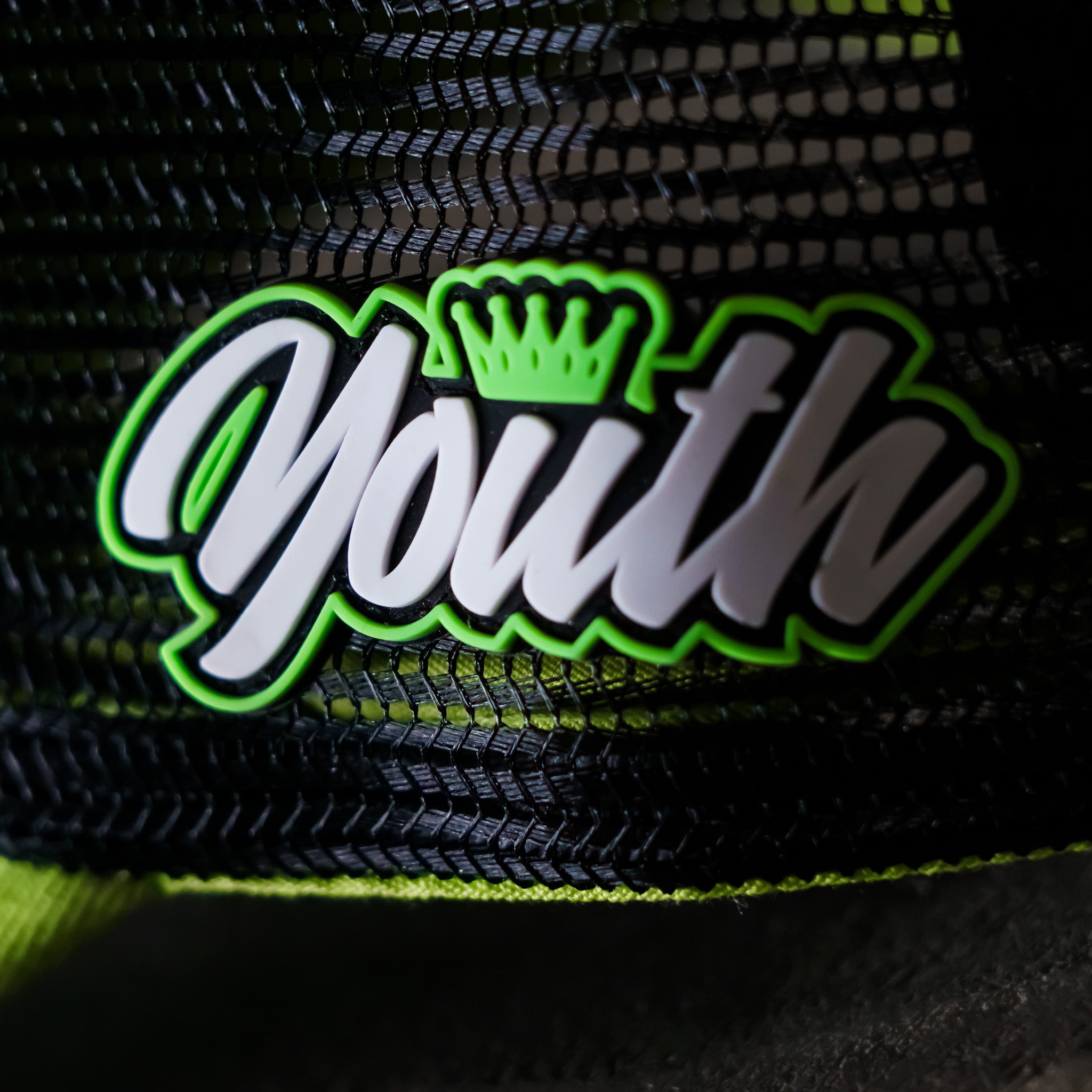 Close-up of the Youth badge on the Uggly & Co Fluro Diamond Trucker Hat x Kyle Ryde, photographed on a concrete floor, highlighting the badge detail on the British Superbikes inspired cap.