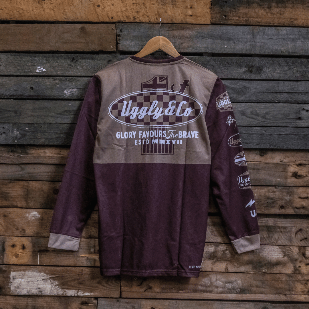 A long-sleeved brown and beige motocross jersey with checkered patterns and text graphics hanging on a wooden background backwards.