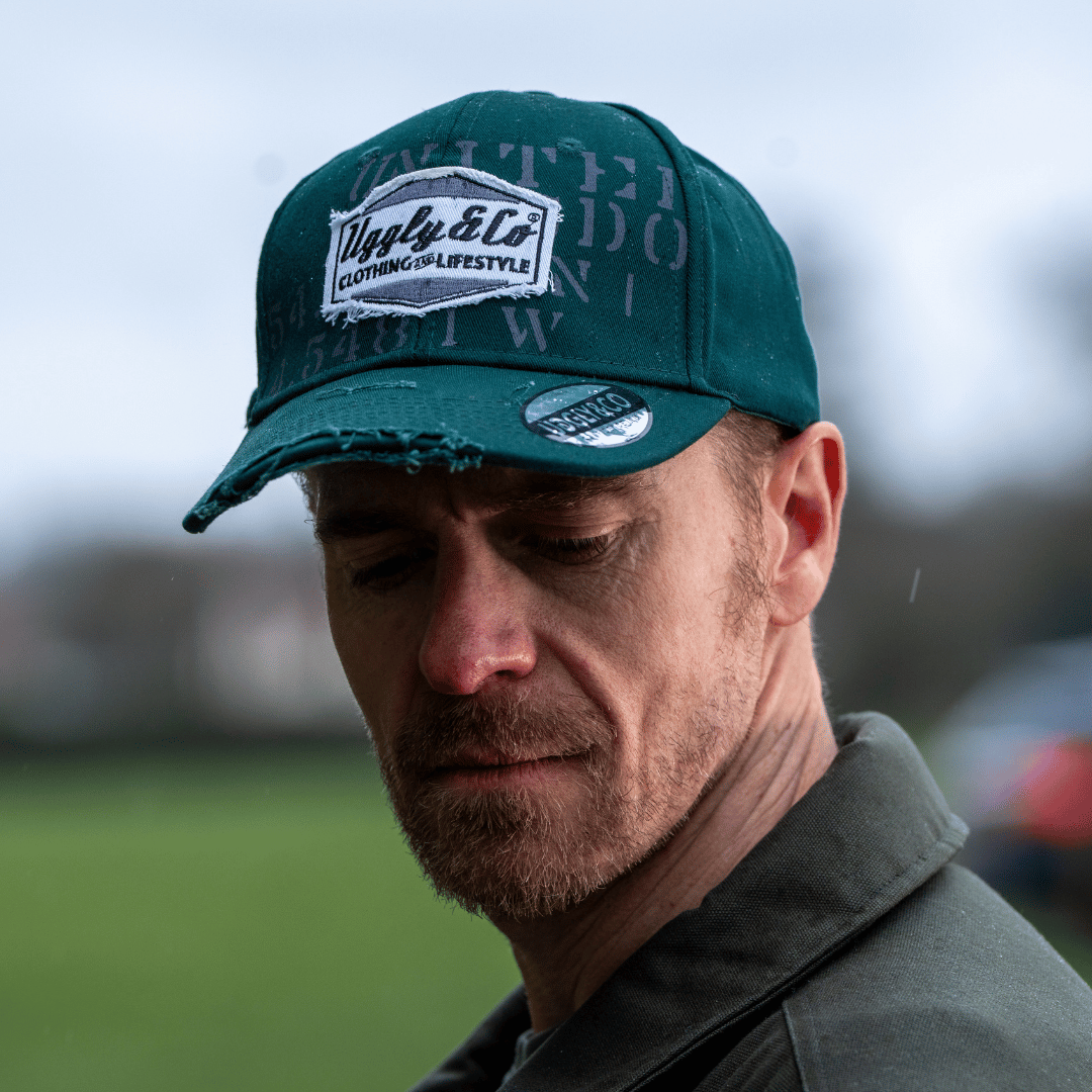 Close up of a man wearing a green 'Aviation' Diamond Trucker Hat with distressed black and white Uggly&Co logo on an airfield