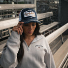 Female model wearing the Uggly&Co Night Blue and Gold Diamond Trucker Hat in a motorsport grandstand