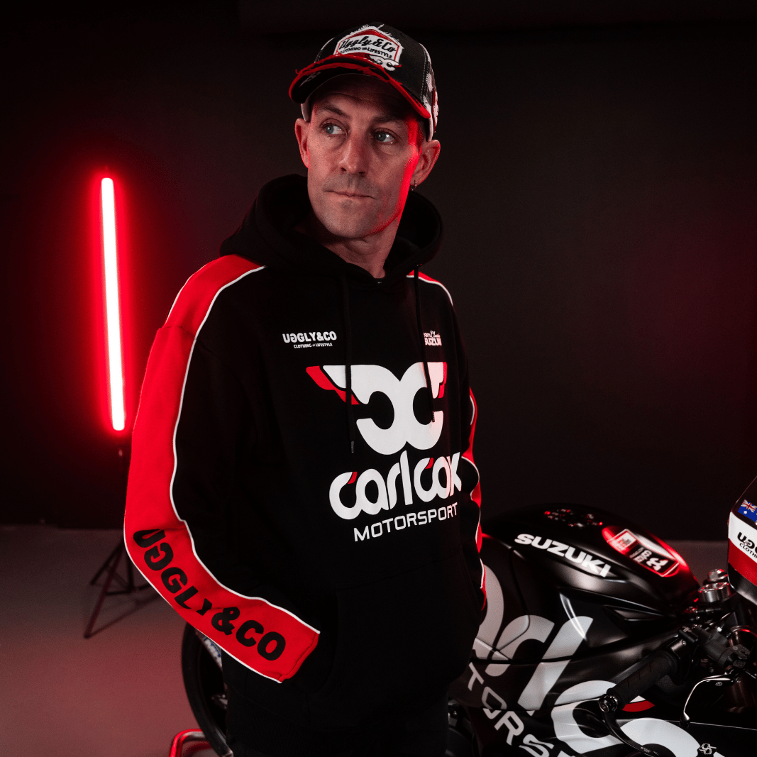 Josh Brookes wearing a black hoodie with red sleeves featuring Carl Cox Motorsport branding in front of his motorbike