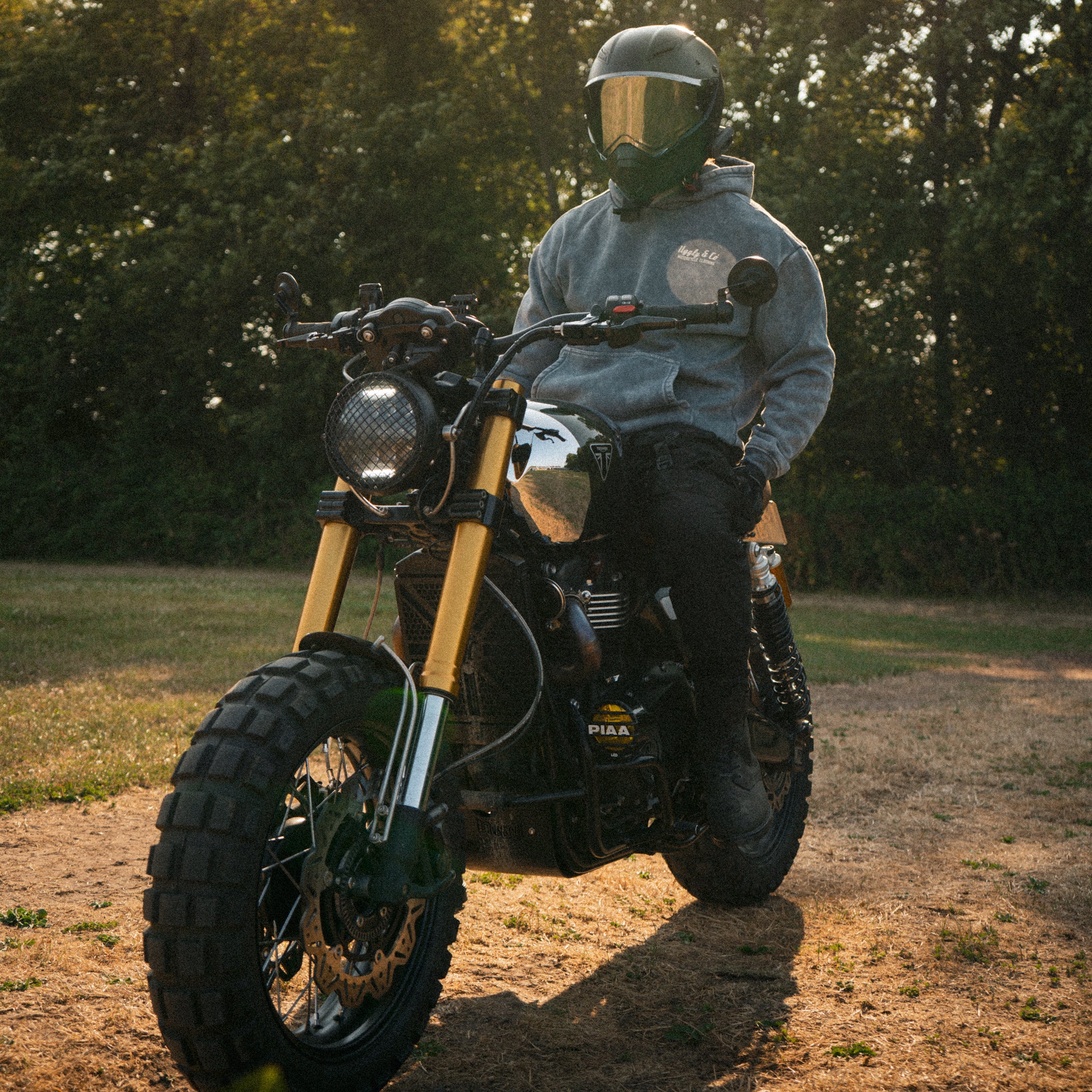 Golden hour lifestyle shot of a male rider in an acid wash hoodie on a Triumph Scrambler bike.