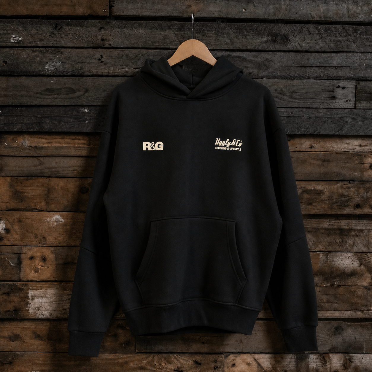 A black pullover hoodie hanging on a wooden hanger against a rustic dark wood plank wall. The hoodie has a kangaroo pocket and two small white chest prints: a block-letter 'FXG' on the wearer's left chest and a script logo on the right chest.