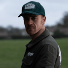 A man wearing a green 'Aviation' Diamond Trucker Hat with distressed black and white Uggly&Co logo on an airfield