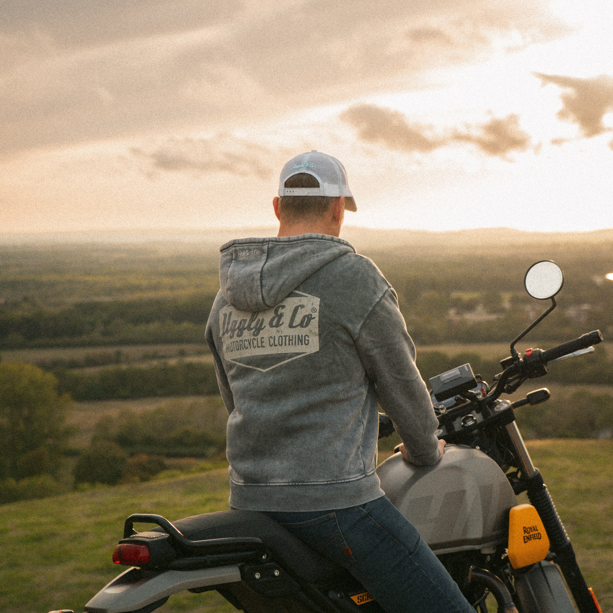 Lifestyle shot of a motorcycle rider in a retro Uggly&Co trucker hat with a distressed peak, beside a Royal Enfield Scram 411