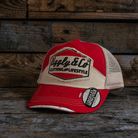 Red and beige trucker hat with Uggly&Co branding and a mesh back section on a wooden background