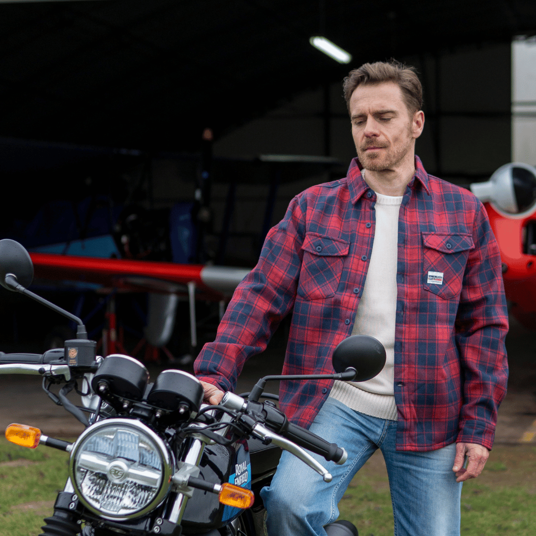 Man in a red plaid shirt standing next to a motorcycle with an airplane in the background.