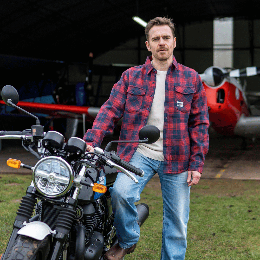 Man in a red plaid shirt standing next to a motorcycle with an airplane in the background.