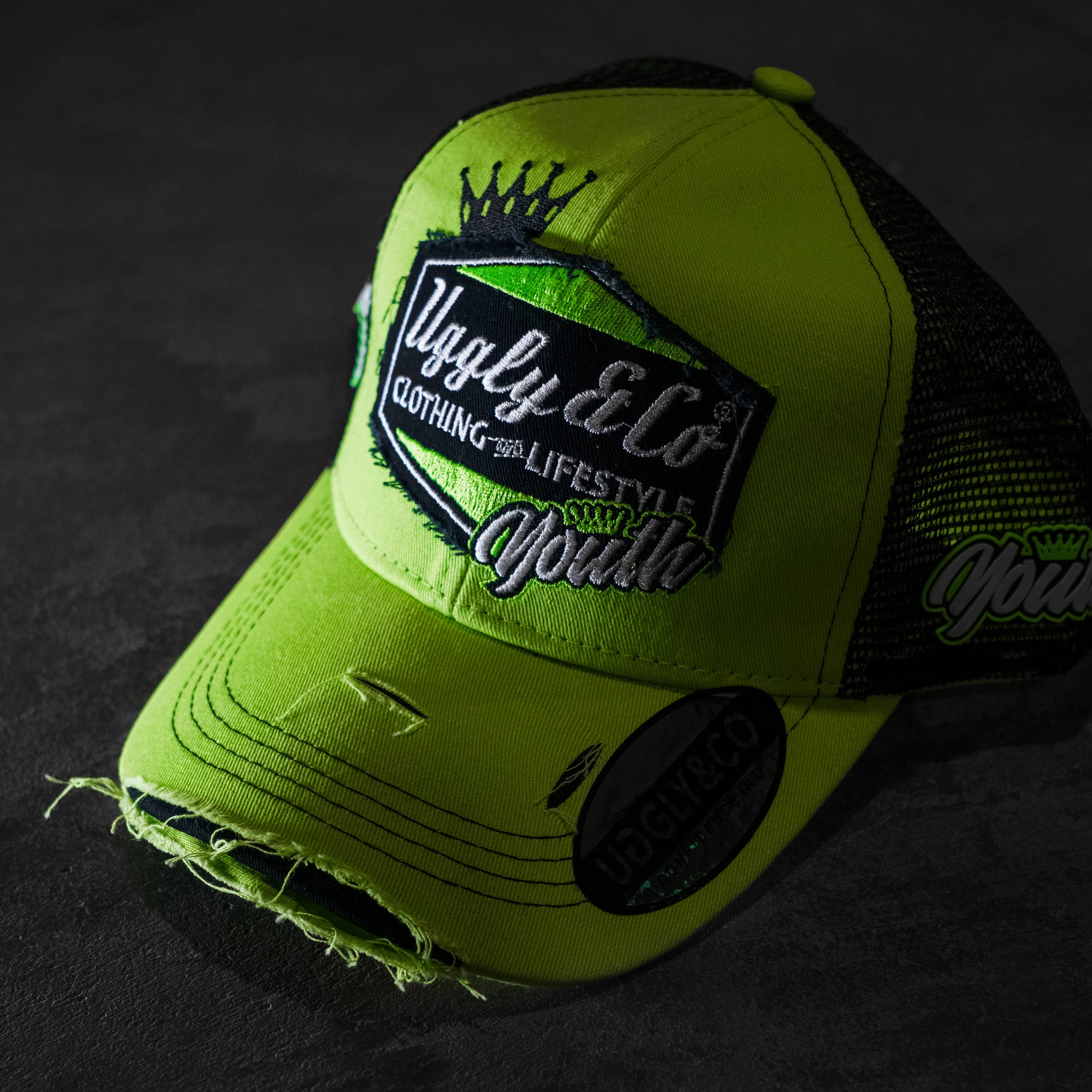 Angled product photo of the Uggly & Co Fluro Diamond Trucker Hat x Kyle Ryde on a concrete floor, highlighting the motorcycle lifestyle cap design.
