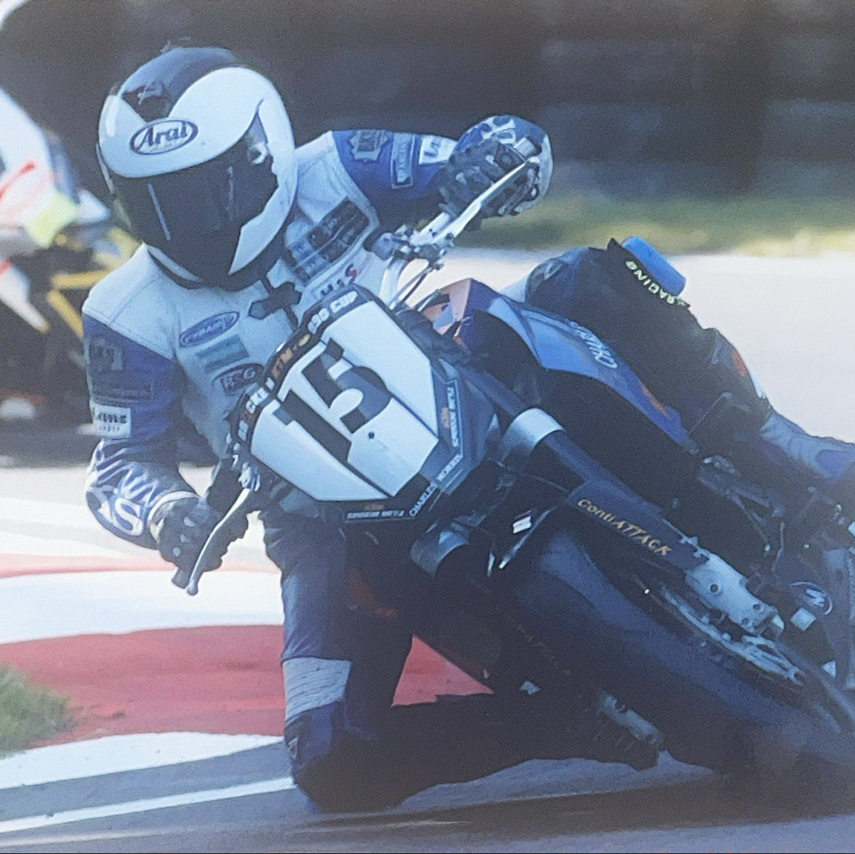 A motorcycle racer leans sharply into a turn on a racetrack, wearing a white and blue racing suit, gloves, and helmet with the Arai logo. The motorcycle is blue and white with the number 15 on the front.