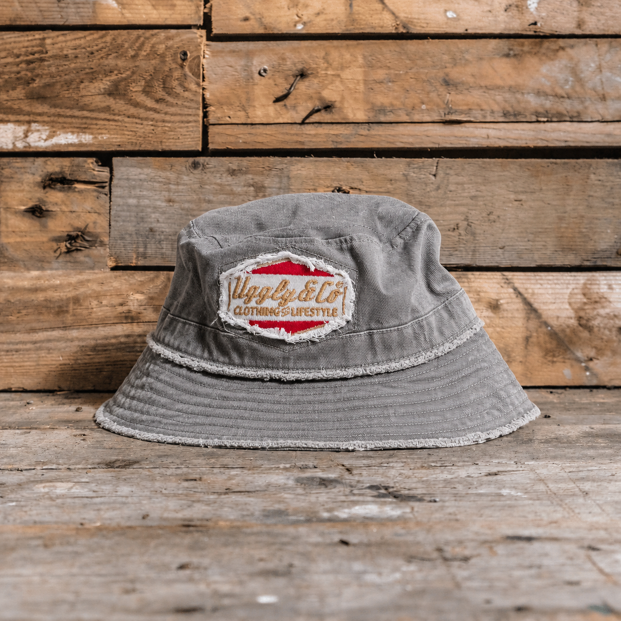 Distressed grey bucket hat with an Uggly&Co diamond logo on a wooden background