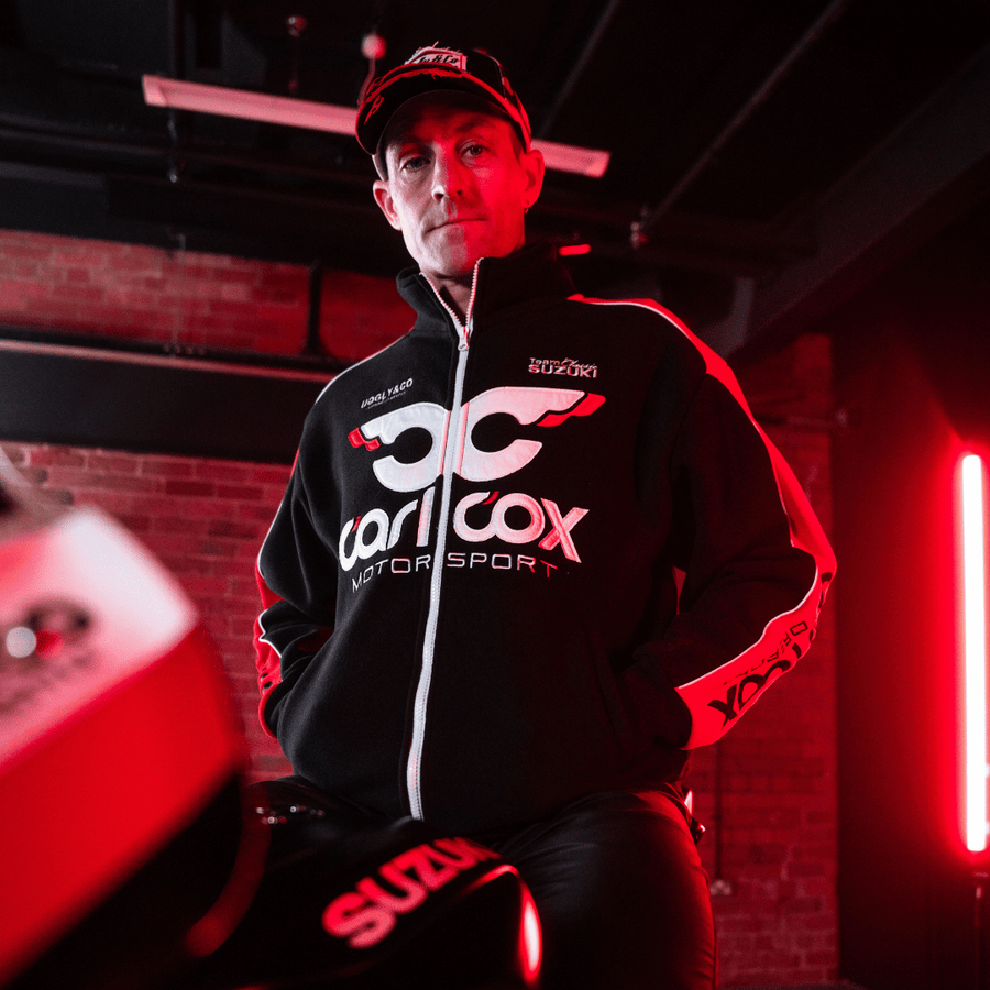 Josh Brookes wearing a black and red jacket with Carl Cox Motor Sport, Uggly&Co and Team Classic Suzuki branding in a blurred garage