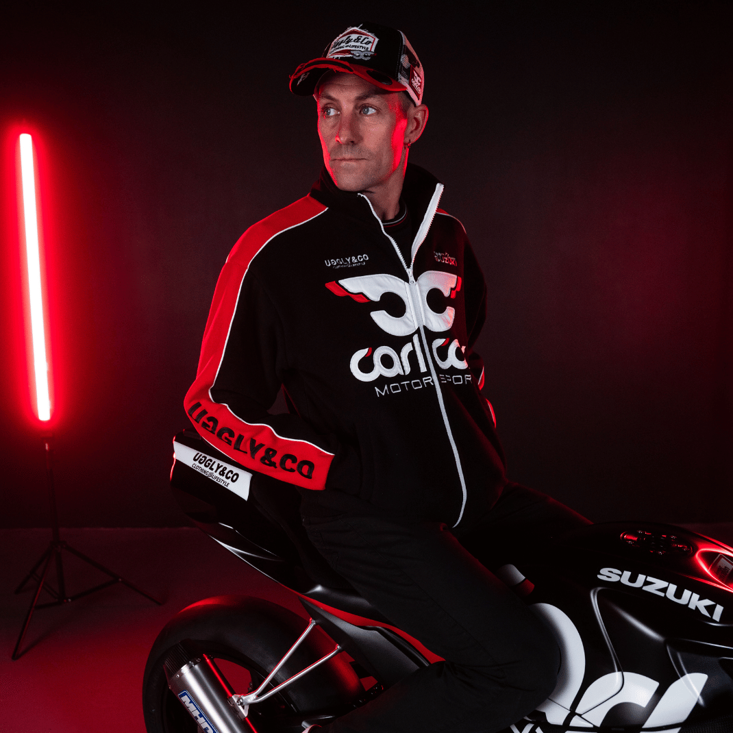 Josh Brookes sitting on his motorbike while wearing a black and red jacket and trucker hat with Carl Cox Motor Sport, Uggly&Co and Team Classic Suzuki branding