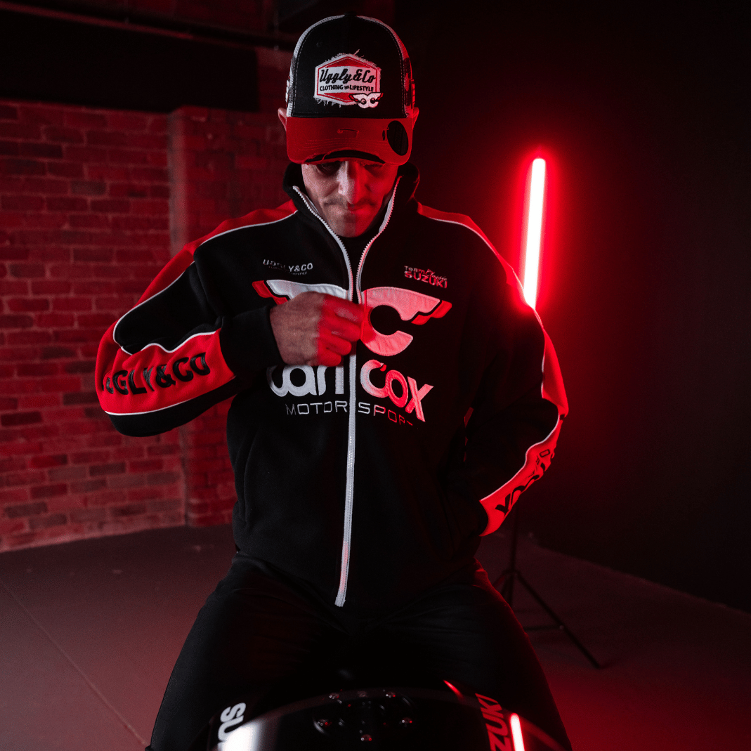 Front view of Josh Brookes sitting on his motorbike while wearing a black and red jacket and trucker hat with Carl Cox Motor Sport, Uggly&Co and Team Classic Suzuki branding