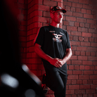 Josh Brookes wearing a Carl Cox motorsport and Team Classic Suzuki black and red t-shirt while leaning against a wall