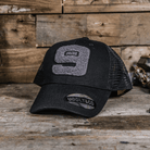 A black trucker cap with the number 9 and the text 'The Black Dub' on the front, laid on a wooden surface.