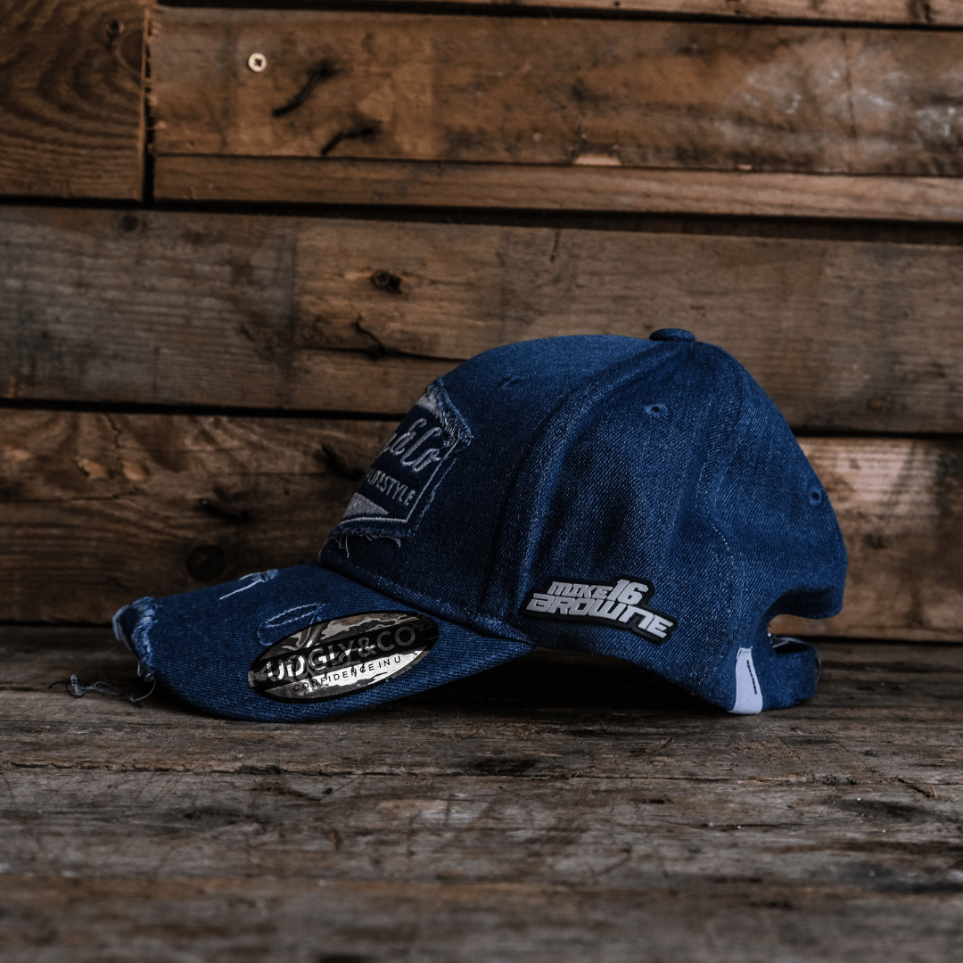 Left side view of a blue denim Baseball Cap with Uggly&Co branding and Mike Browne 16 logo on the side on a wooden background
