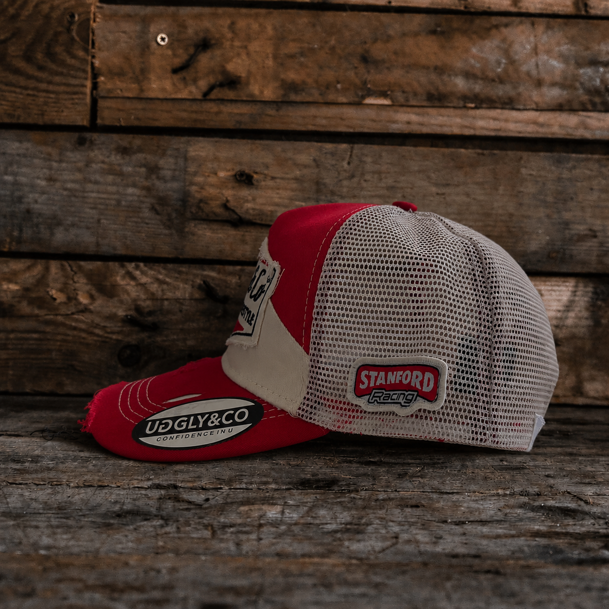 Side view of a red and beige trucker hat with Uggly&Co branding and a mesh back section on a wooden background