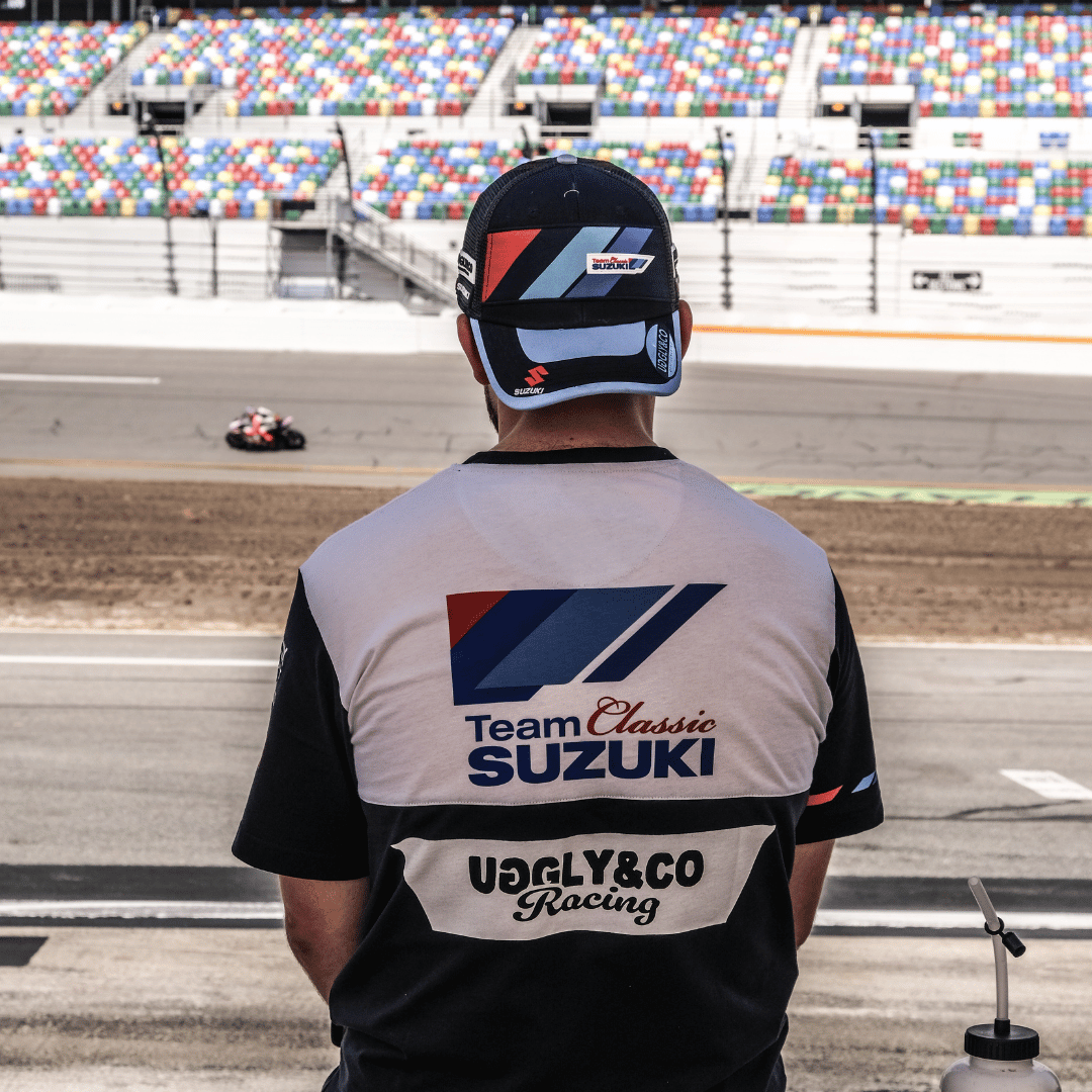 Rear view of a model wearing a trucker hat backwards with geometric pattern featuring blue, red, and black colours, with the Team Classic Suzuki and Suzuki logos on it by a race track