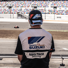 Rear view of a model wearing a trucker hat backwards with geometric pattern featuring blue, red, and black colours, with the Team Classic Suzuki and Suzuki logos on it by a race track