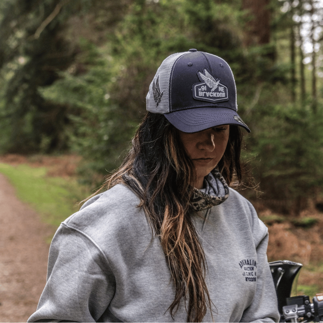 A woman wearing a dark navy blue trucker hat with a grey mesh back featuring a Black Dub Garage Spark Plug logo on the front in the woods