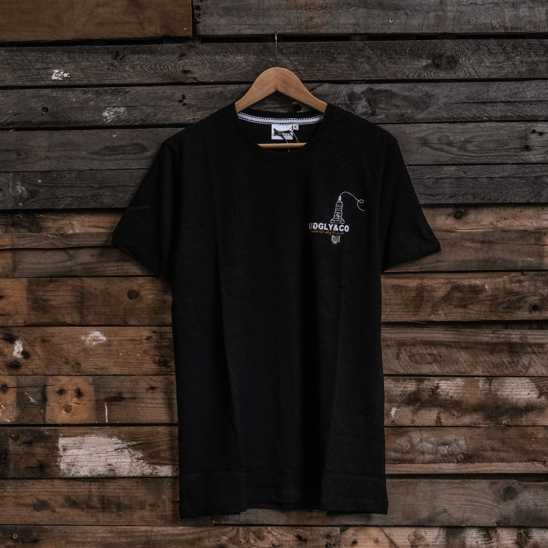 Front view of a black t-shirt with a spark plug graphic and the Uggly&Co logo on a wooden background